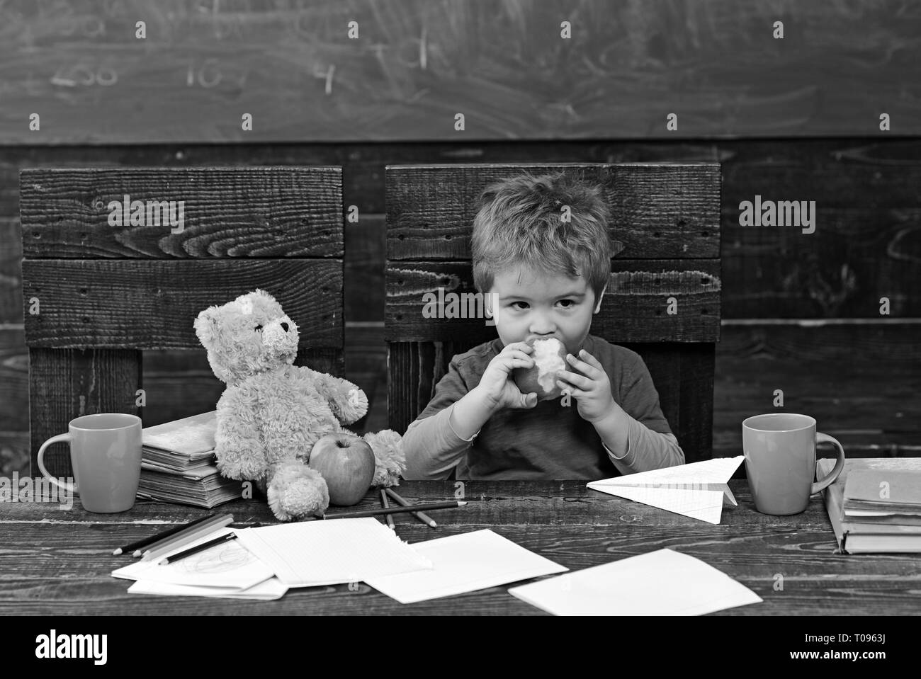School break. Hungry kid biting apple in classroom. Small boy playing with paper plane and teddy bear Stock Photo