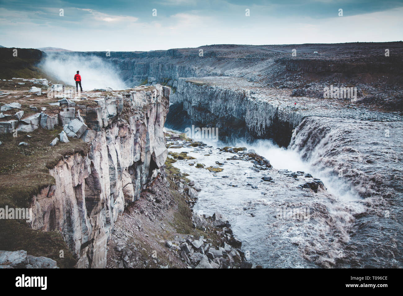 Panorama view of hiker in red jacket standing in front of gigantic Dettifoss waterfall on a moody day with dark clouds in summer, Skogar, Iceland Stock Photo