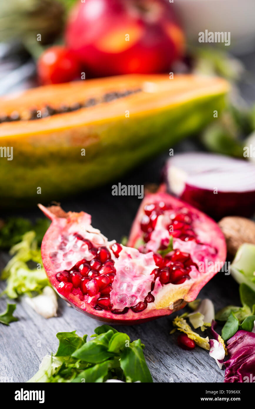 closeup of a halved pomegranate on a gray rustic wooden table full of different fruits and vegetables, such as half papaya, chopped lettuce, a purple  Stock Photo