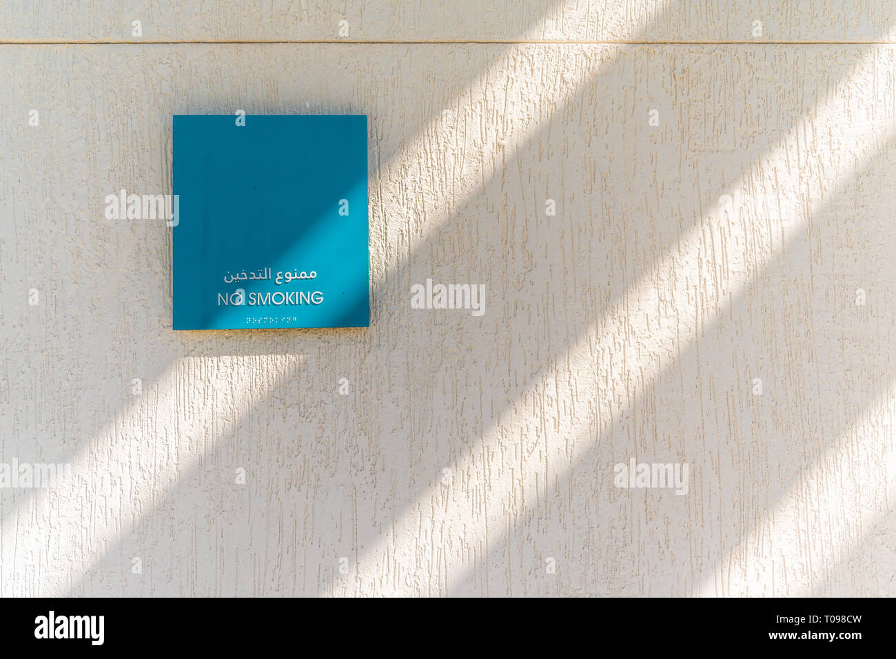 A blue no smoking sign on textured white wall and rays of sunlight, Umm Al Emarat Park, Abu Dhabi, United Arab Emirates Stock Photo