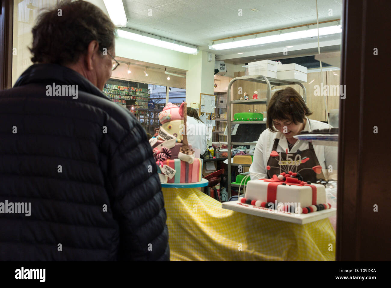 A visitor looks through a shop window as a confectioner decorates a cake at the undercover Golden Cross Market in  Oxford, Oxfordshire, Britain Stock Photo