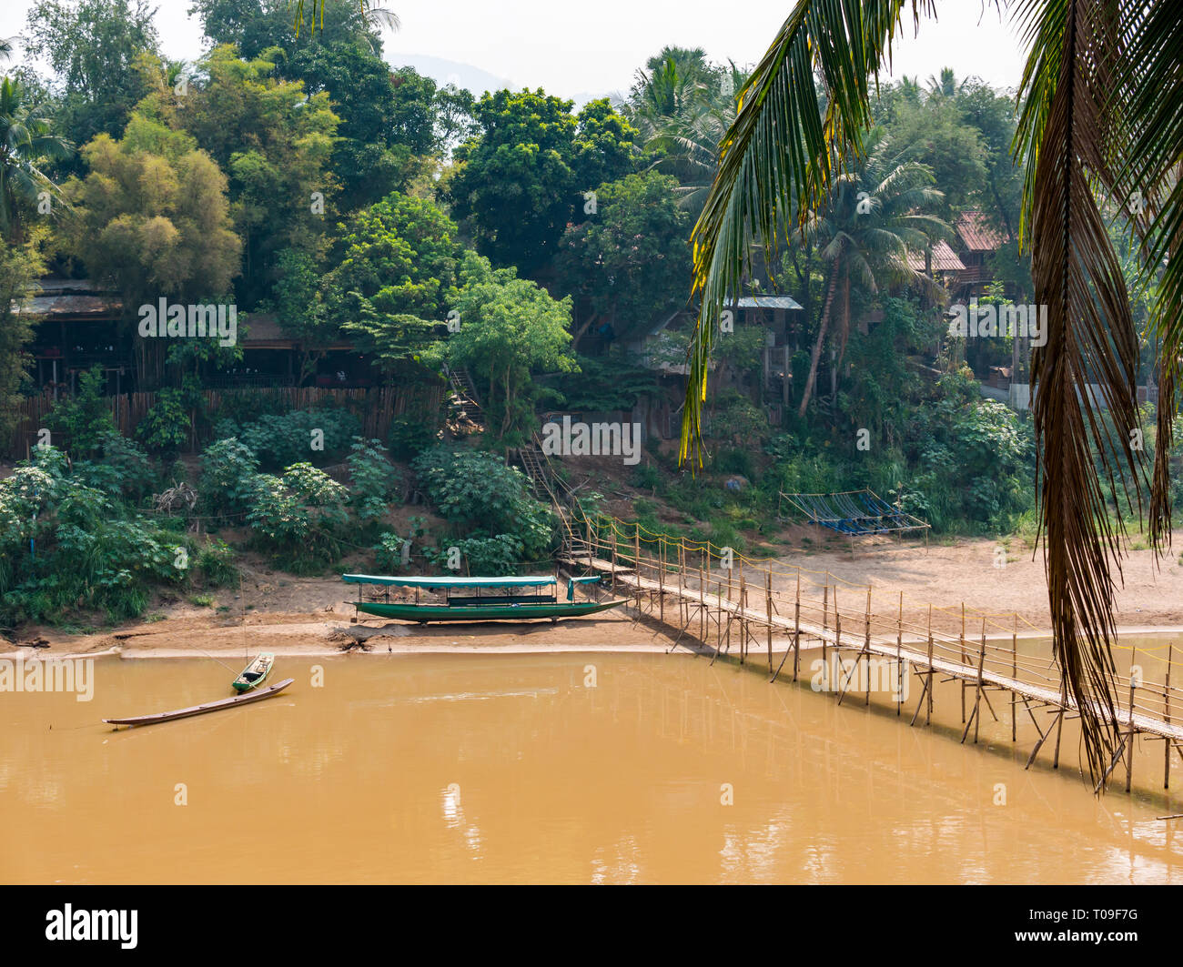 Bamboo cane bridge over Nam Kahn river tributary of Mekong with traditional canopied dugout canoe riverboat, Luang Prabang, Laos, Indochina, SE Asia Stock Photo