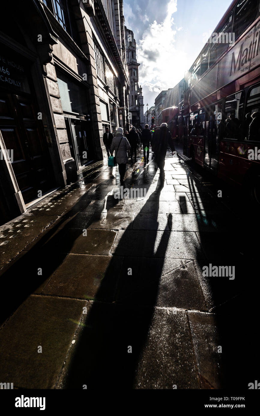 Long shadows on wet pavement as people walk towards low sun along Piccadilly, London, UK. Winter light. Late afternoon shadow. Red London bus Stock Photo
