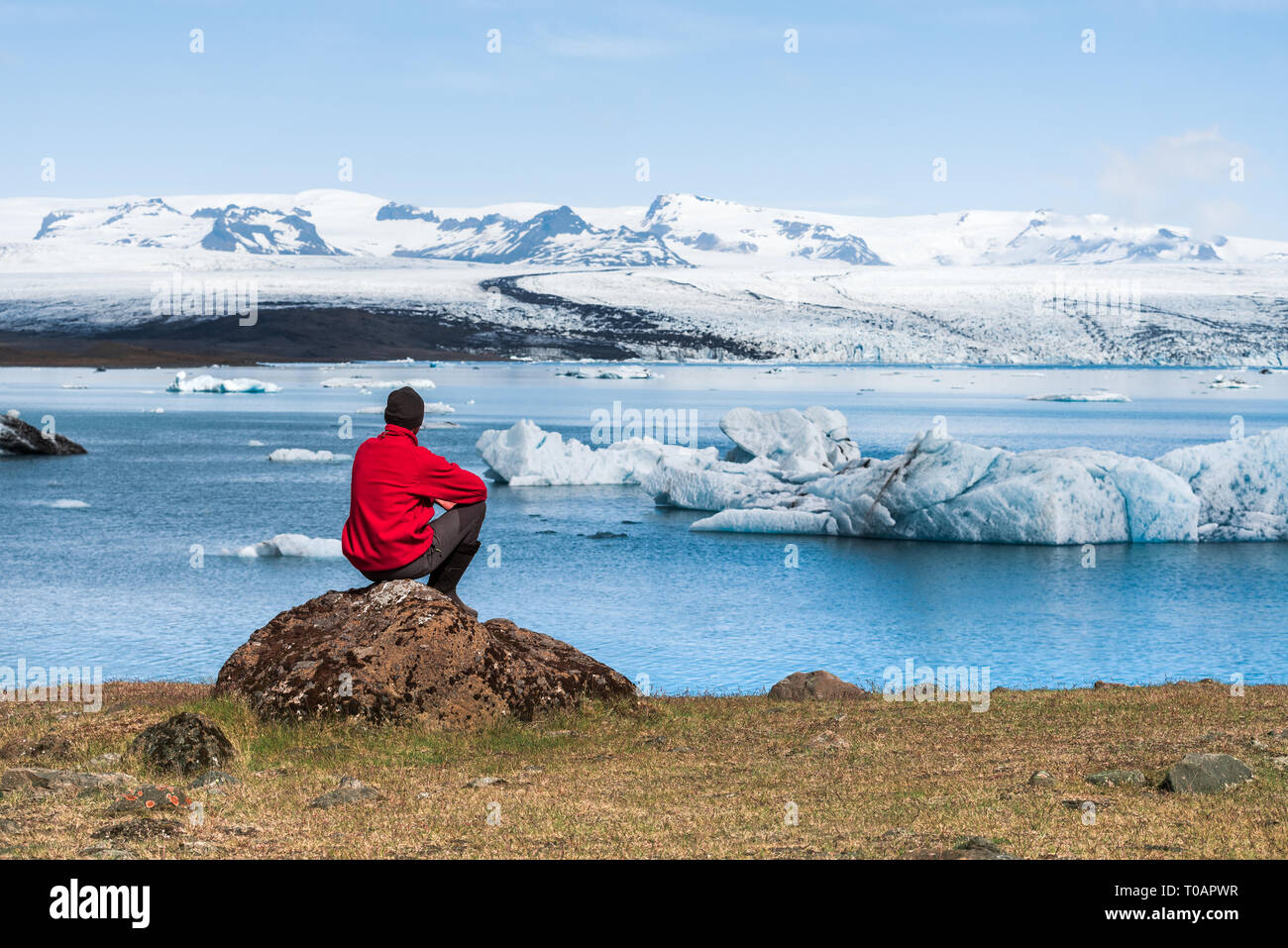Man traveler in red jacket sits on the shore of Jokulsarlon Glacial Lagoon. Summer landscape, glacier and icebergs of Iceland. National park Vatnajoku Stock Photo