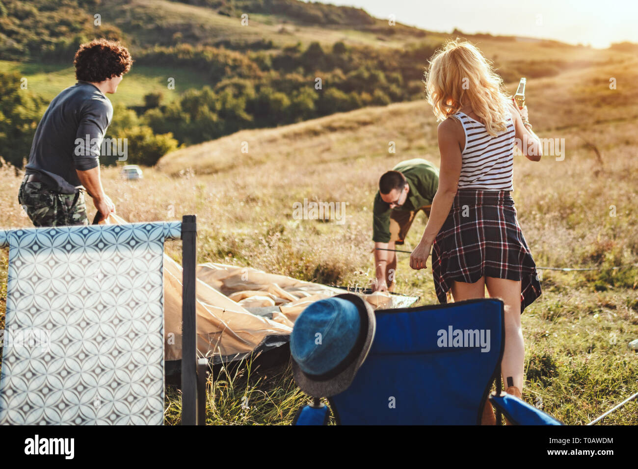 The young happy friends are preparing for camping. They're installing a tent on a suitable place in a meadow and their girlfriends are offering the be Stock Photo