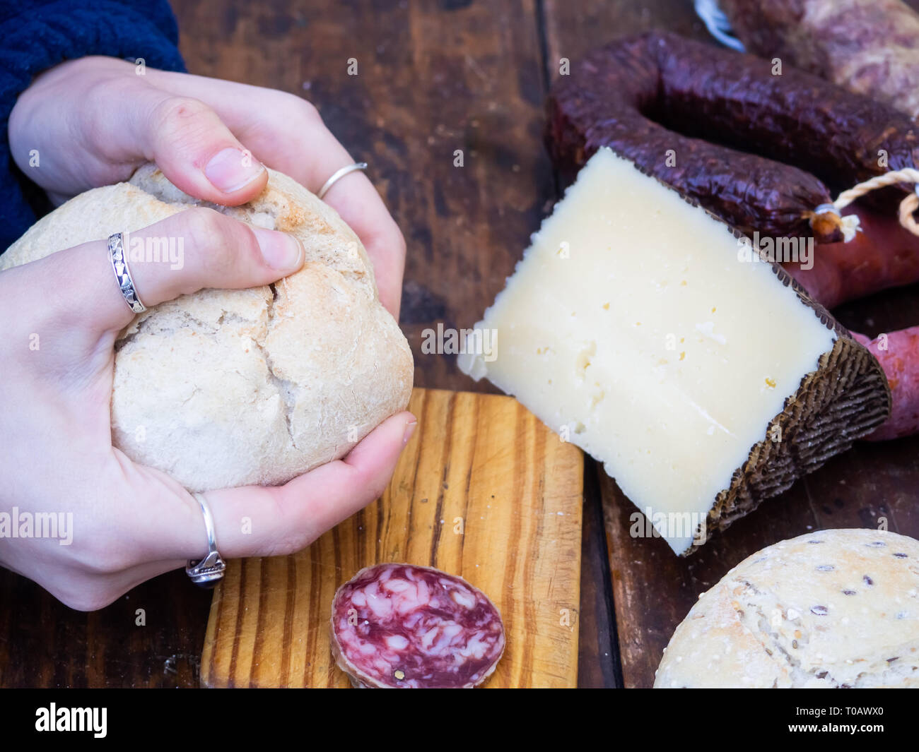 One person cutting rustic bread and Iberian pork sausage and a wedge of sheep cheese on an old wooden board Stock Photo