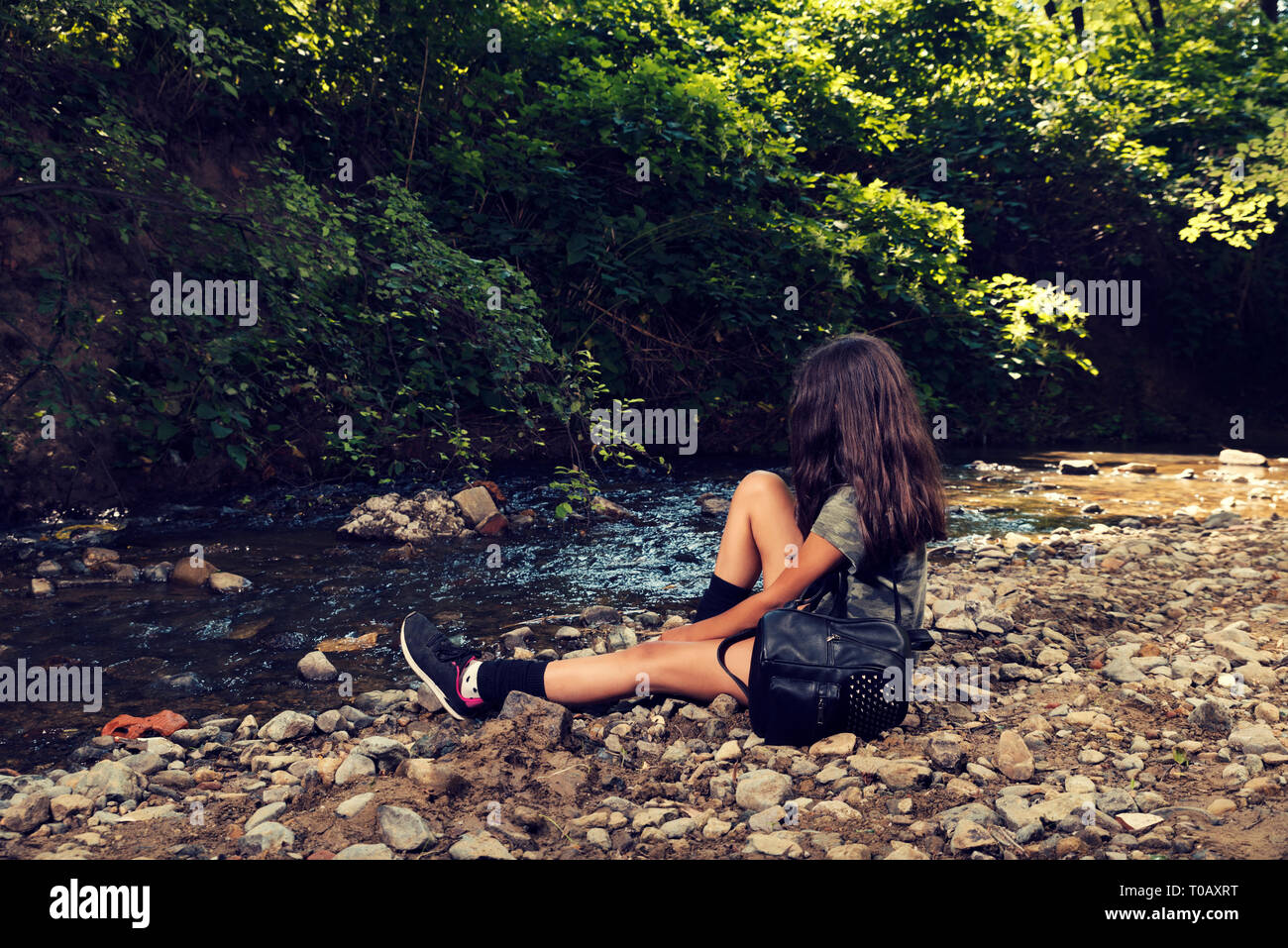 Young girl with black backpack sitting alone on on a river bank  in a city park Summer time Selective focus Stock Photo