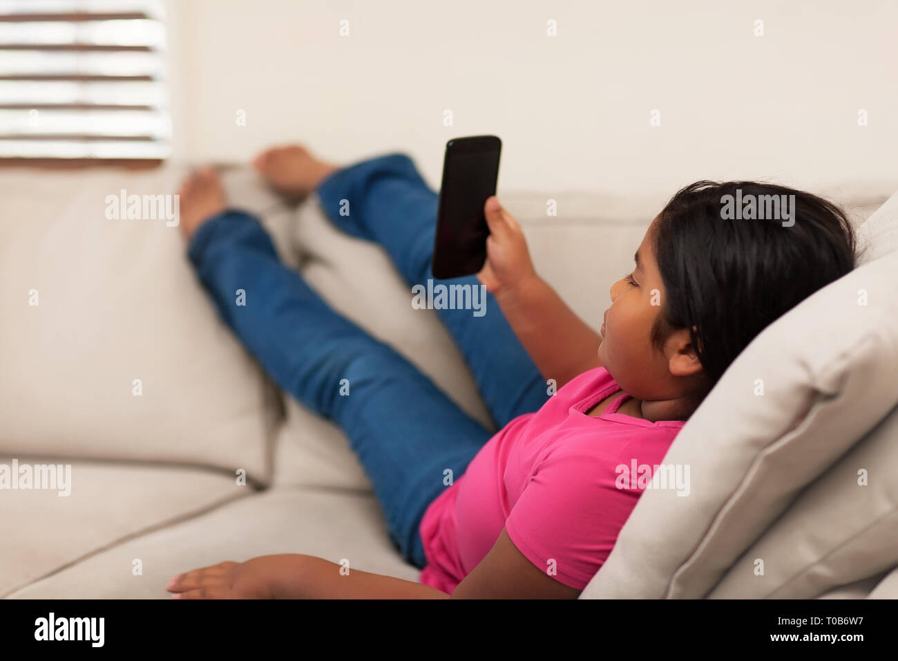 Mixed race 8 year old girl watching mobile phone while stretching out her legs; sitting on a couch with a relaxed pose. Stock Photo