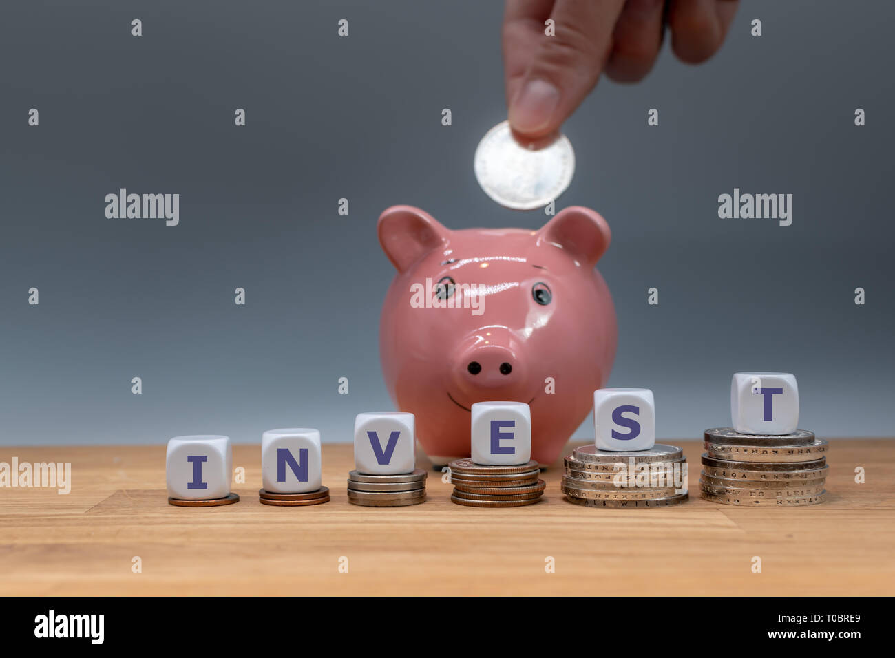 Dice form the word 'INVEST'. Dice placed on stacks of coins. A coin is put into a piggy bank in the background. Stock Photo