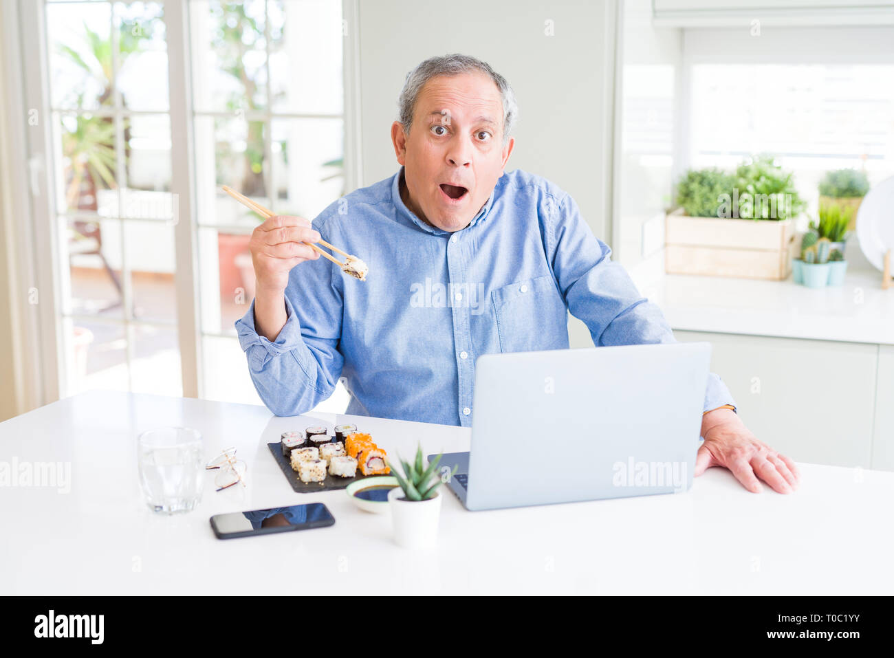Handsome business senior man eating delivery sushi while working using laptop scared in shock with a surprise face, afraid and excited with fear expre Stock Photo