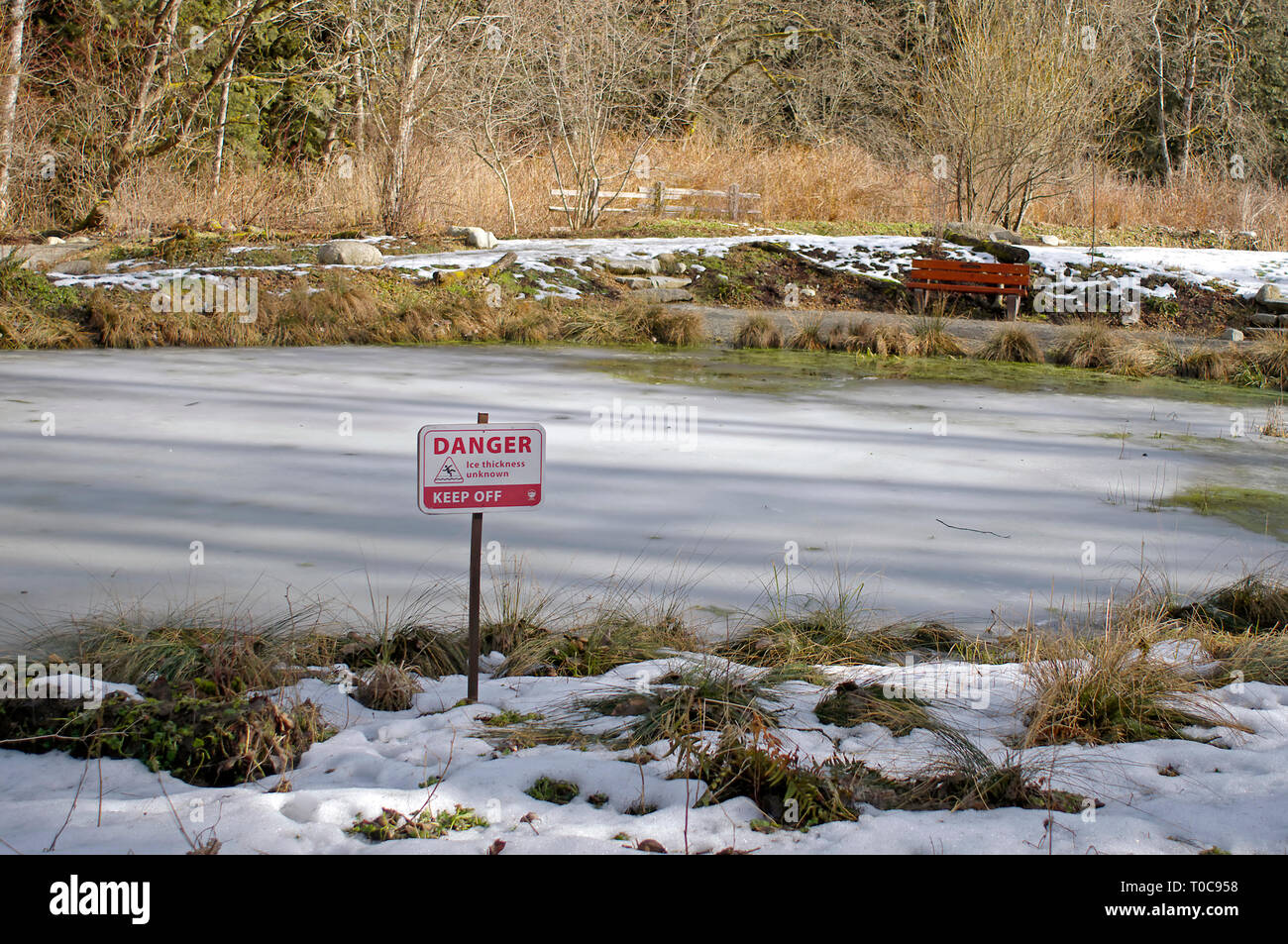 Warning sign reading 'Danger  Ice Thickness Unknown Keep Off' beside an ice-covered pond with a bench on the far side. Stock Photo