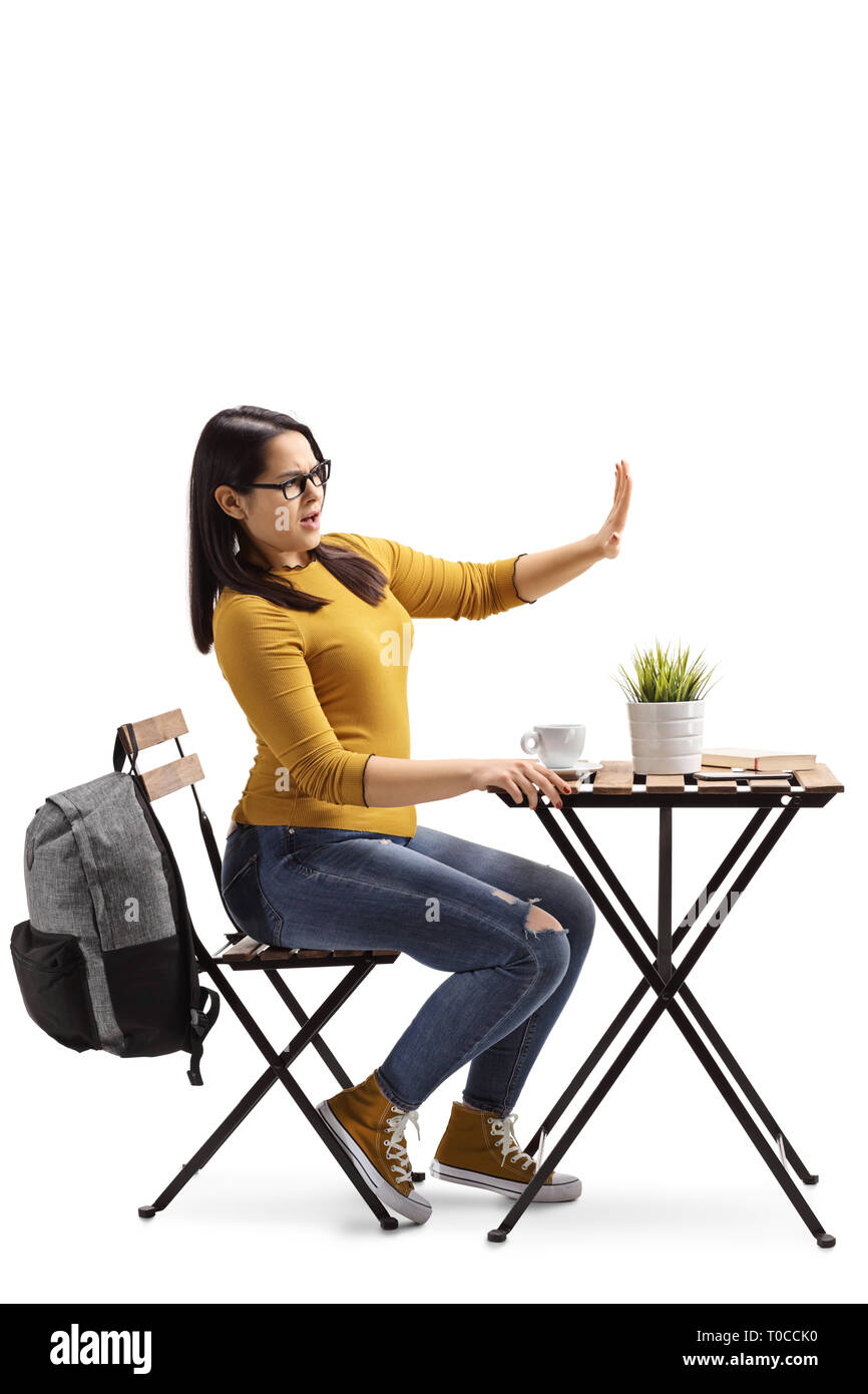 Full length profile shot of an angry female student in a cafe sitting at a table with coffee and gesturing stop with hand isolated on white background Stock Photo