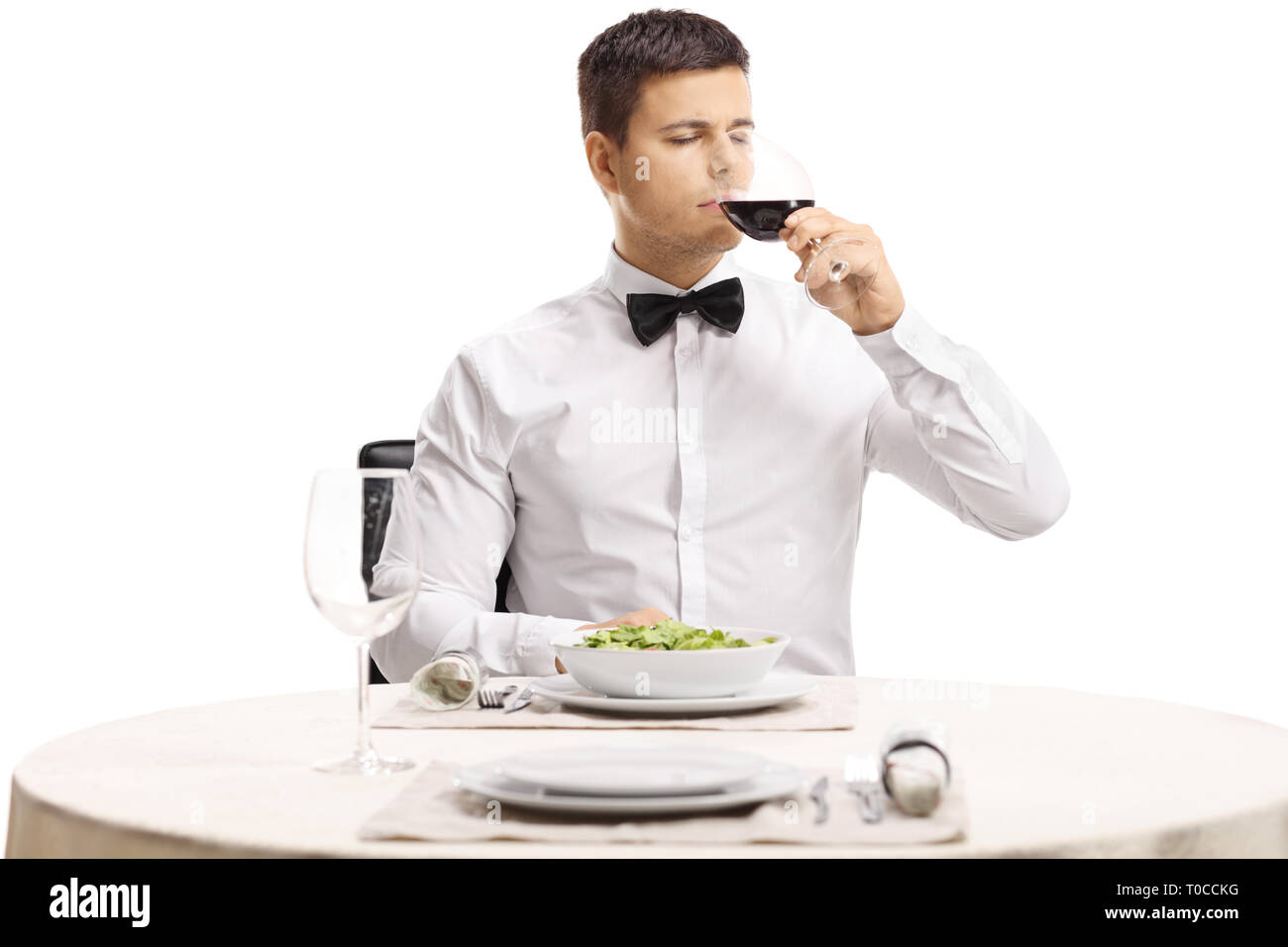 Portrait of a young handsome man with a bow tie tasting red wine at a restaurant table isolated on white background Stock Photo