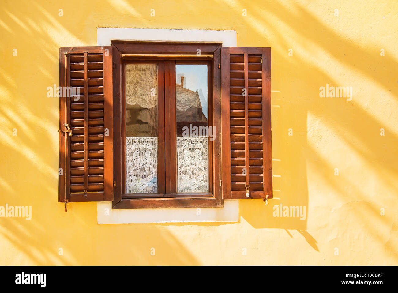 beautiful window with wooden shutters in yellow wall on bright sunny summer day with tree shadow on the wall Stock Photo