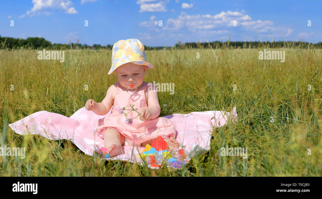 Cute little girl in a hat sitting on pink blanket, green field and blue sky in the background Stock Photo