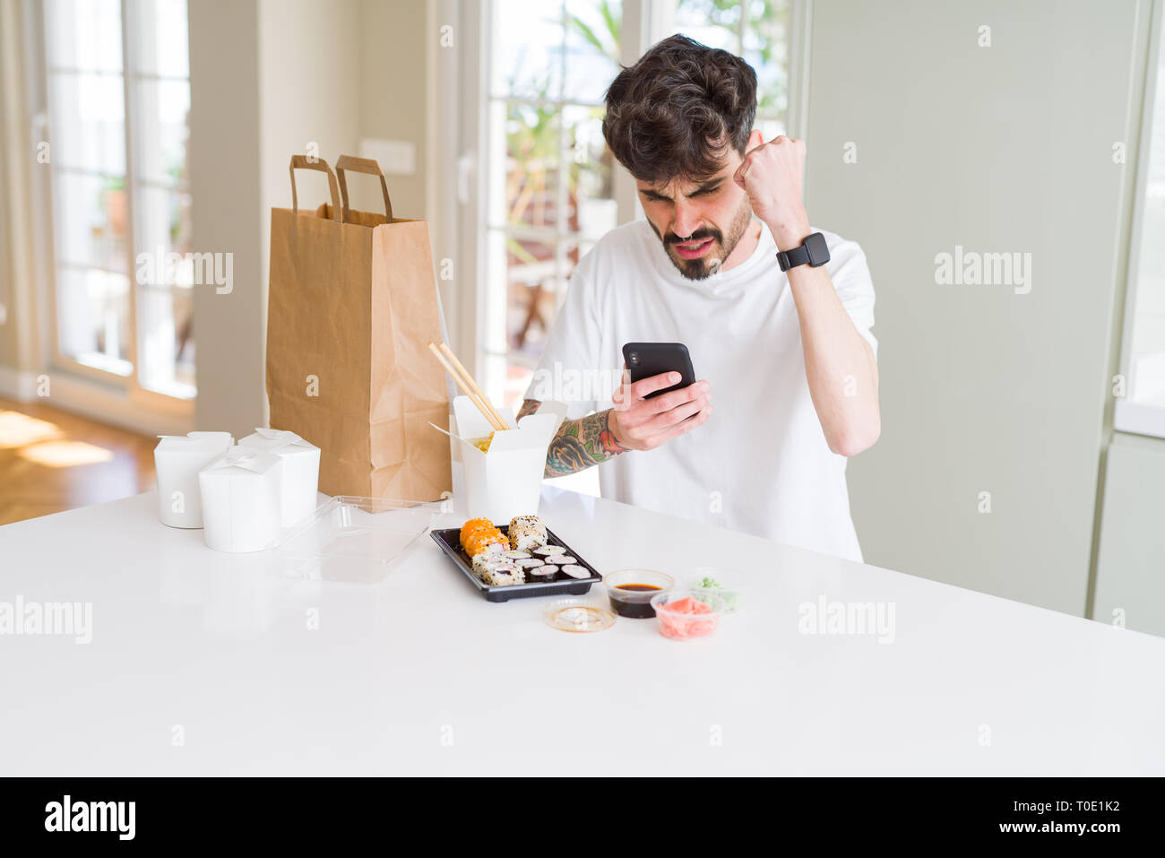 Young man eating asian sushi from home delivery and ordering food using smartphone app annoyed and frustrated shouting with anger, crazy and yelling w Stock Photo