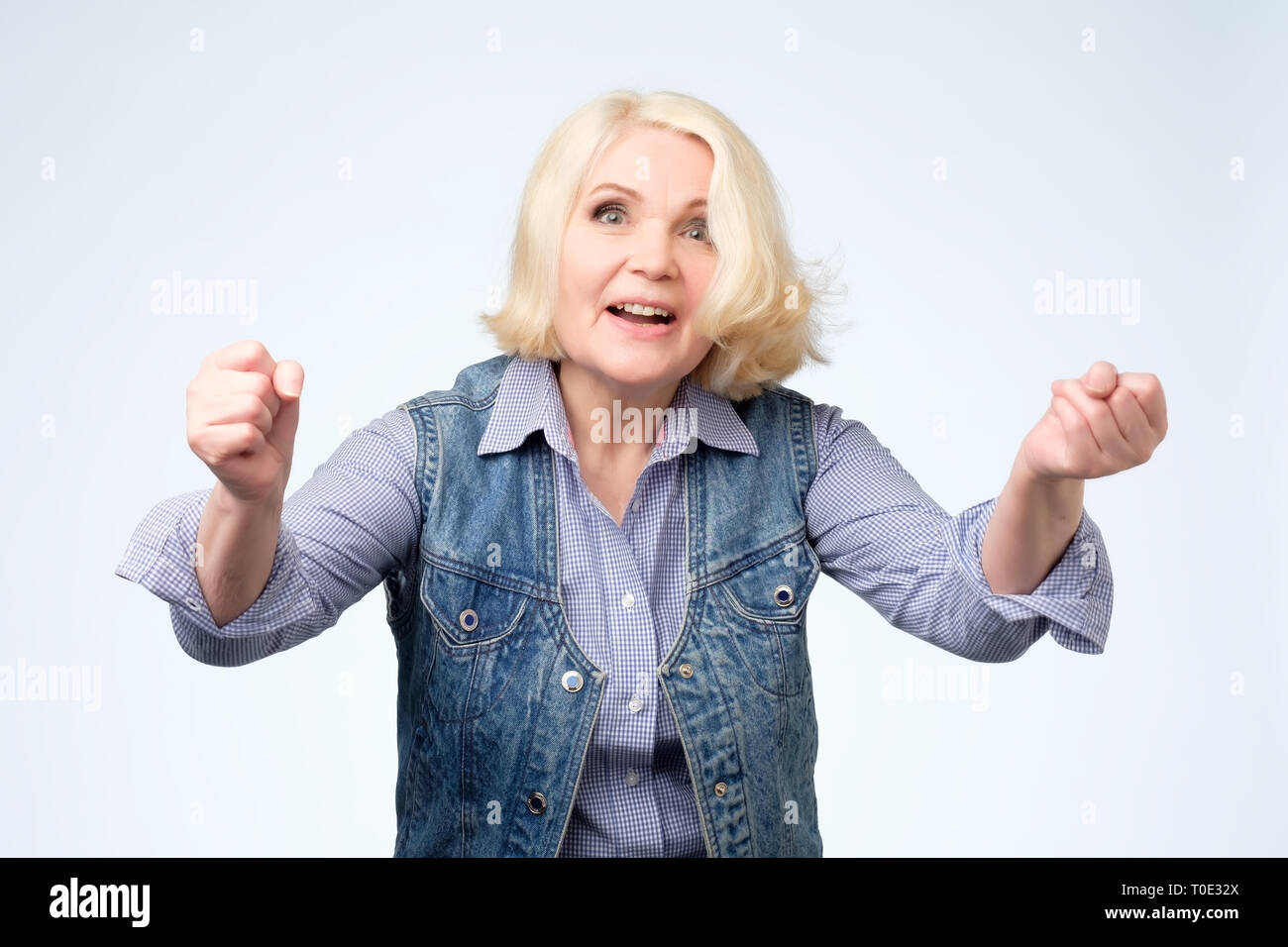 Cheerful granny with toothy smile raised hands and showing successful sign Stock Photo