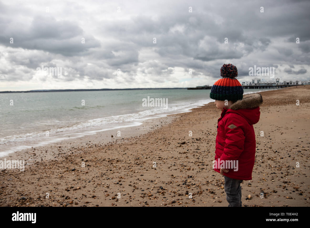 Young boy aged two in red coat playing on the beach in spring with bobble hat Stock Photo