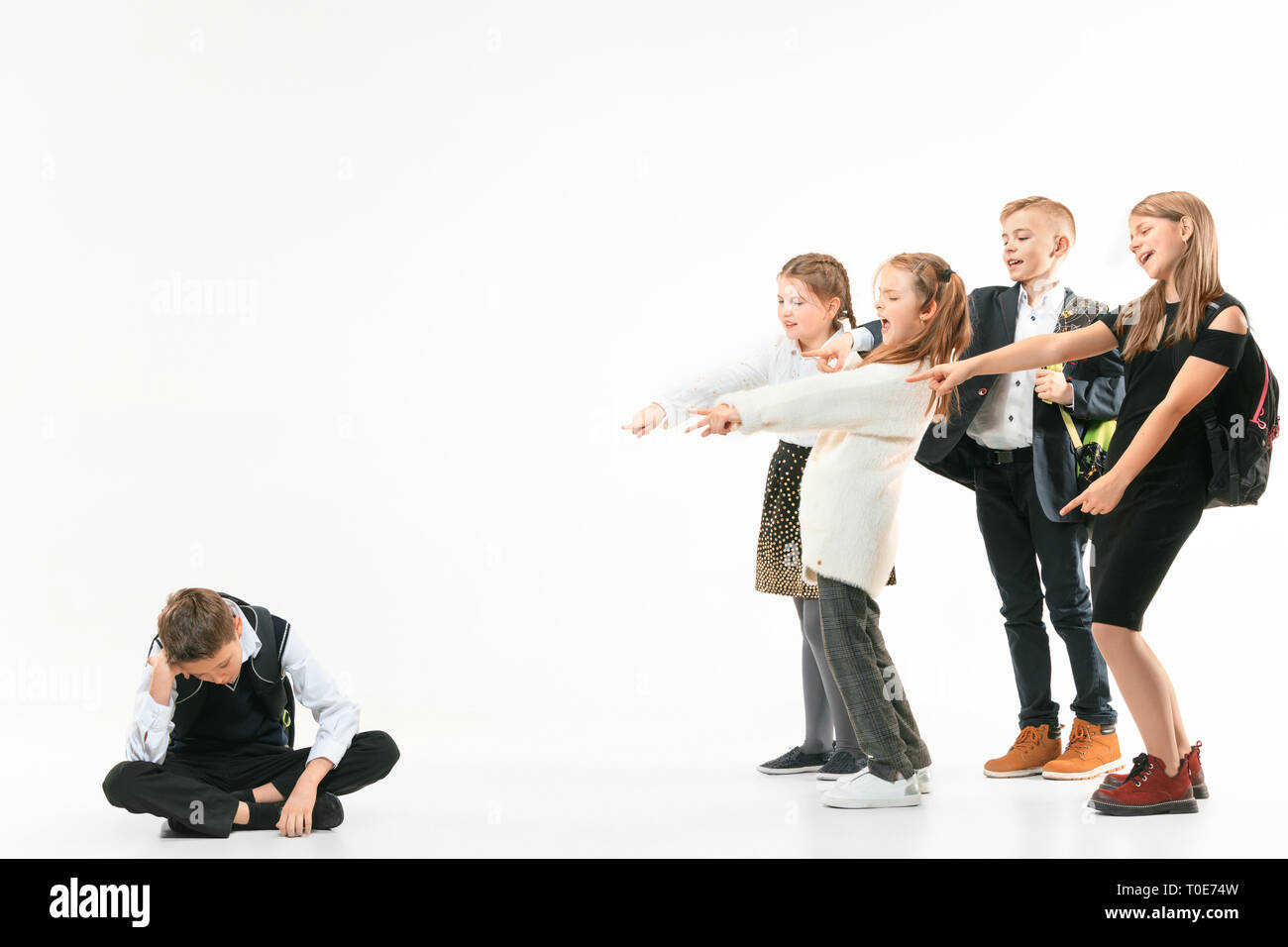 Little boy sitting alone on floor and suffering an act of bullying while children mocking in the background. Sad young schoolboy sitting on studio against white background. Stock Photo