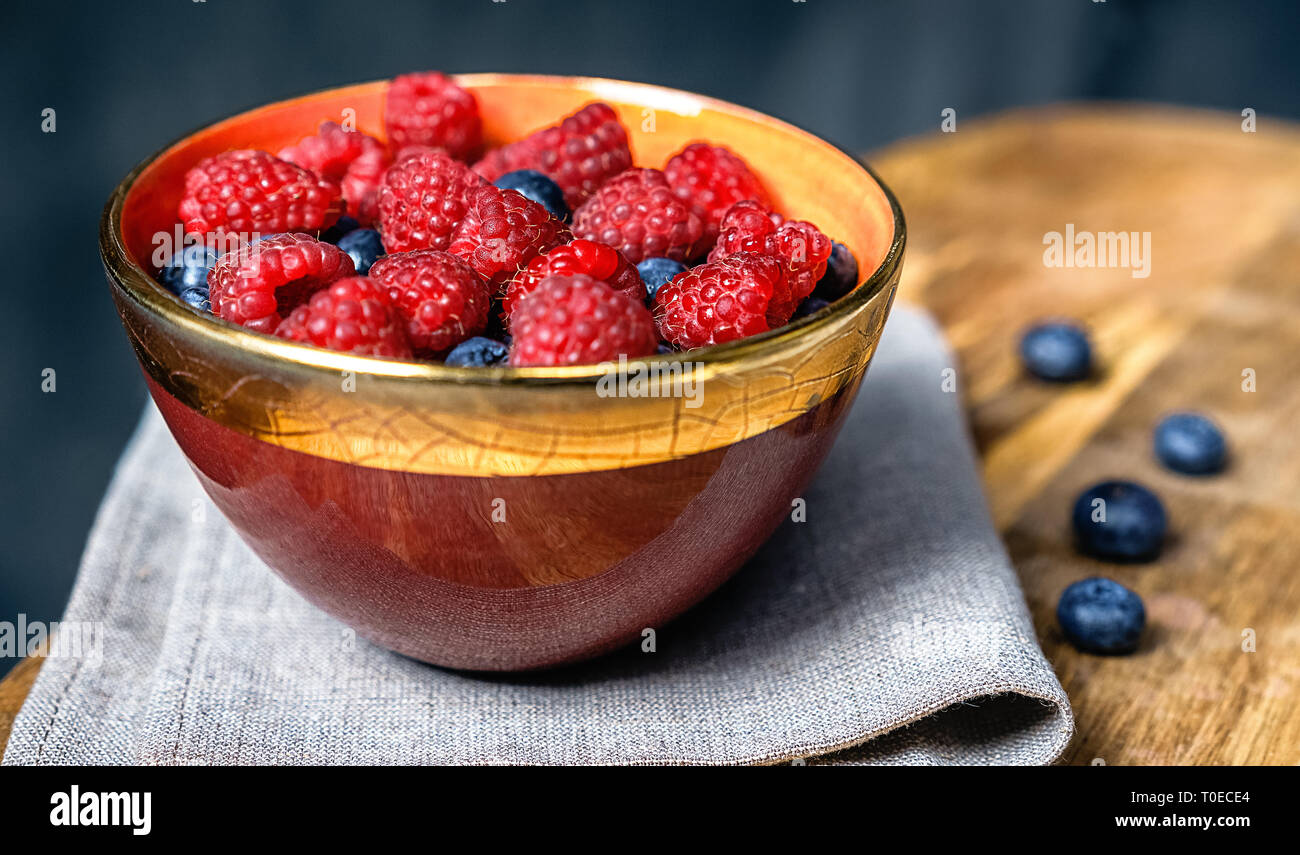 Ripe sweet raspberries and blueberries in bright ceramic bowl on gray tabble cloth on wooden kitchen board. Close up, top view, healthy breakfast ingredient Stock Photo