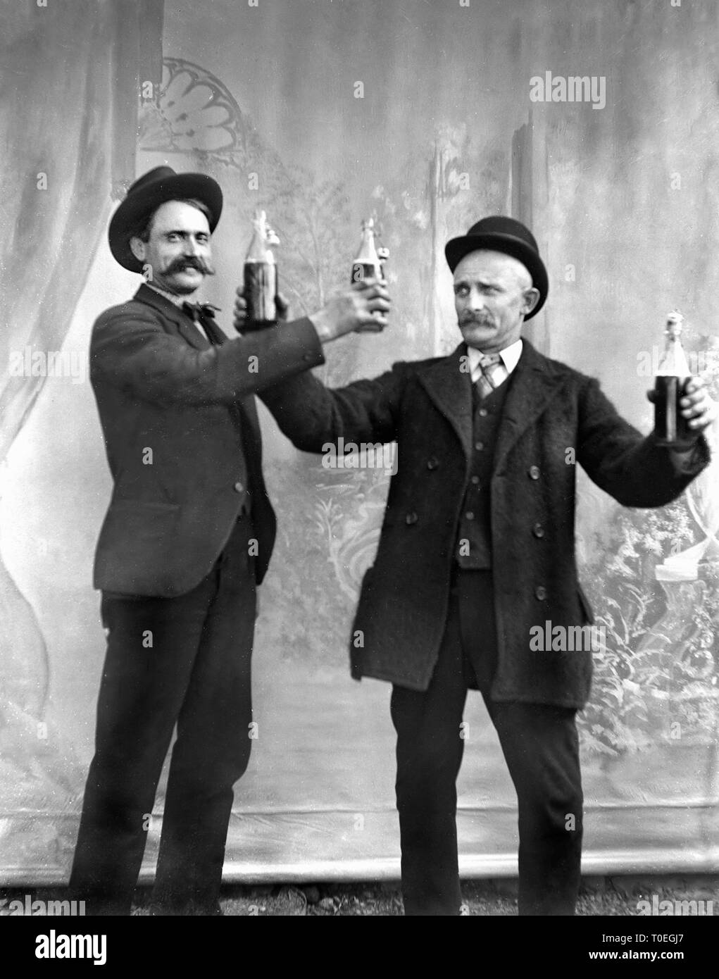 Two early twentieth century men toast each other with beer bottles for a studio portrait, ca. 1905. Stock Photo