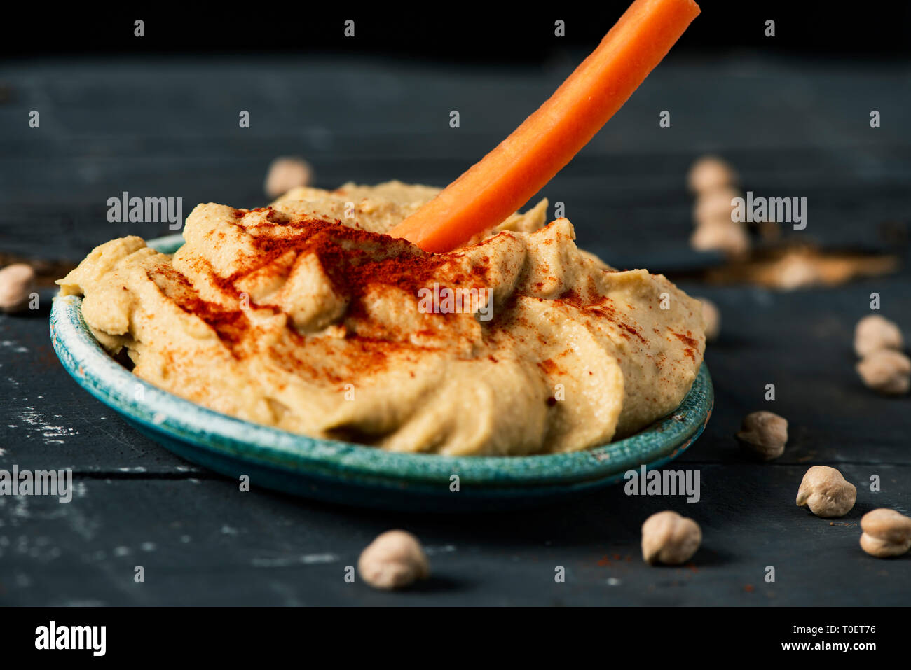 closeup of a strip of carrot being dipped in a homemade hummus seasoned with paprika served in a green ceramic plate, on a dark gray rustic wooden tab Stock Photo