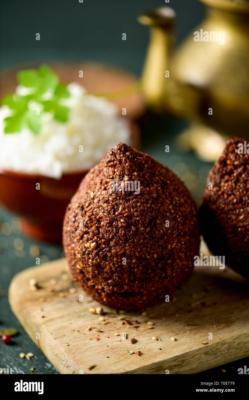 some kibbeh, a levantine dish, made of bulgur, onion, minced meat and different spices, on a rustic wooden table, with a bowl with rice and a golden t Stock Photo