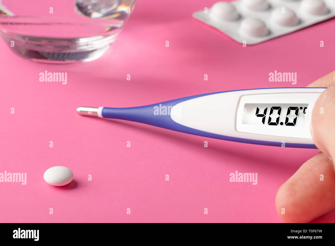 Electronic thermometer and pills on a pink background. High temperature 40 degrees Celsius on display. Glass cup with water. Copy space. Stock Photo