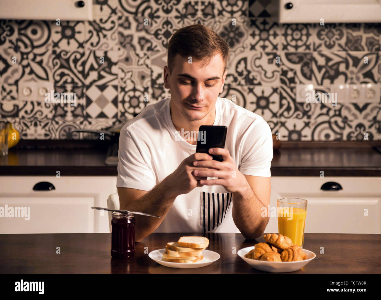 Attractive young man in pajama texting on his smartphone while having breakfast at home Stock Photo