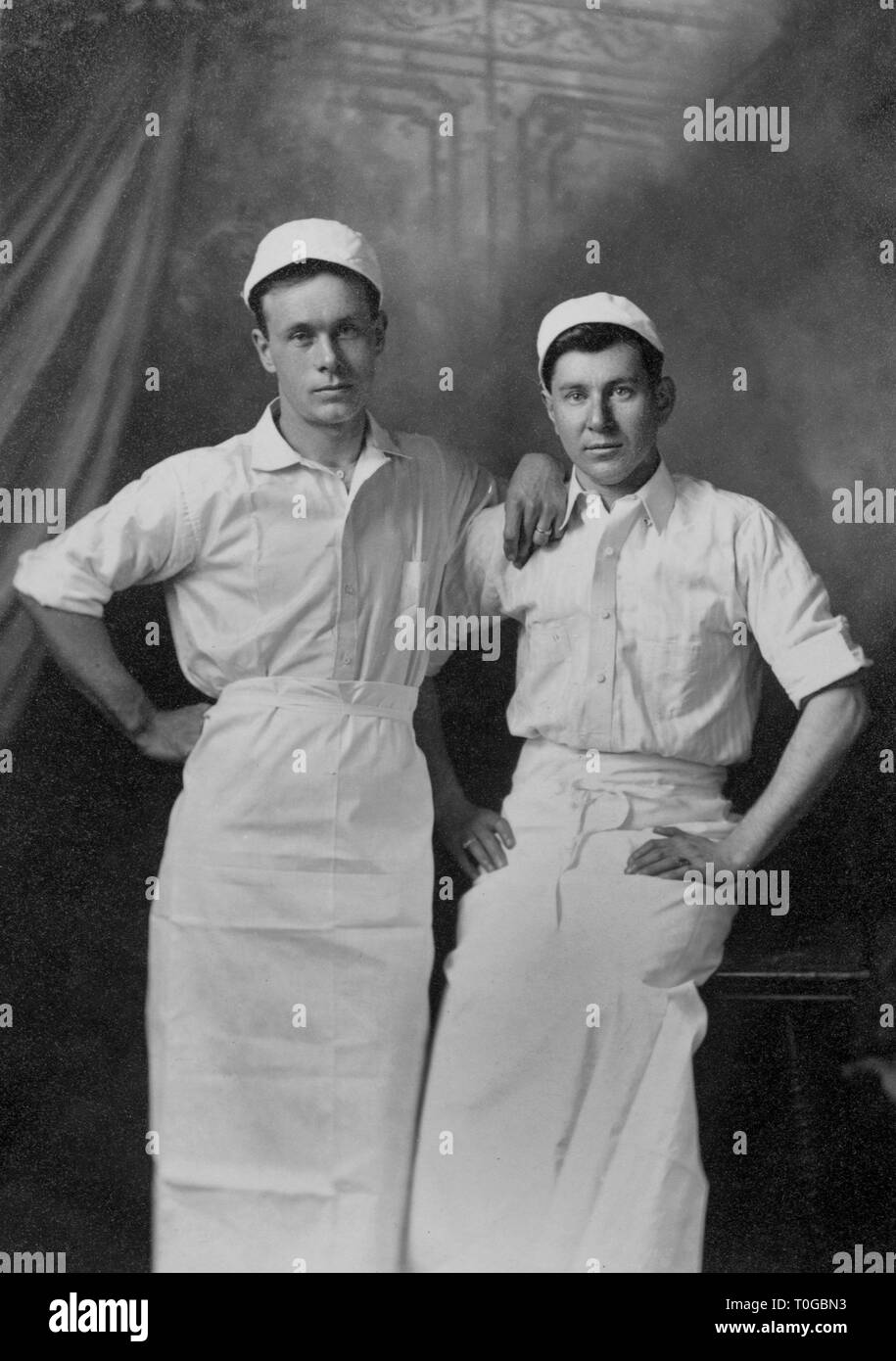 Two food industry workers, possibly bakers or butchers, pose together for a studio portrait, ca. 1910. Stock Photo