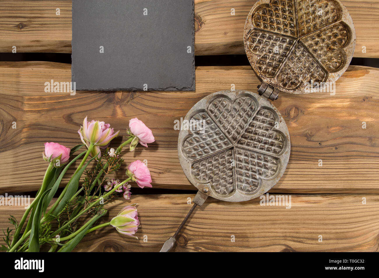 Traditional ancient waffle iron arranged on rustic wood planks with slate board and bunch of flowers. Shot from above, flat lay. Stock Photo