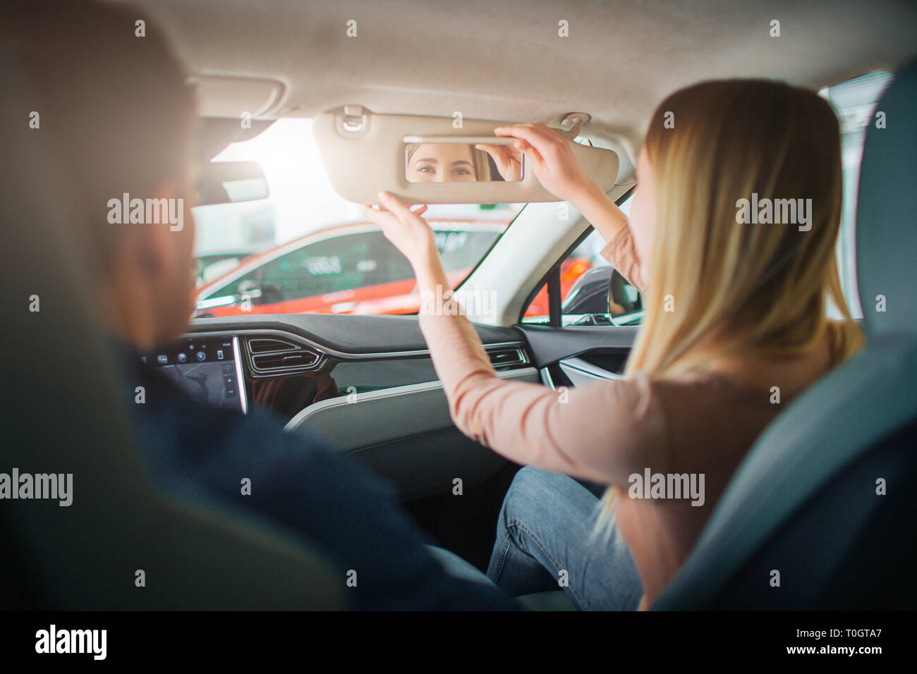Young family buying first electric car in the showroom. Attractive woman looking into the mirror in the cabin of modern electric hybrid vehicle before Stock Photo