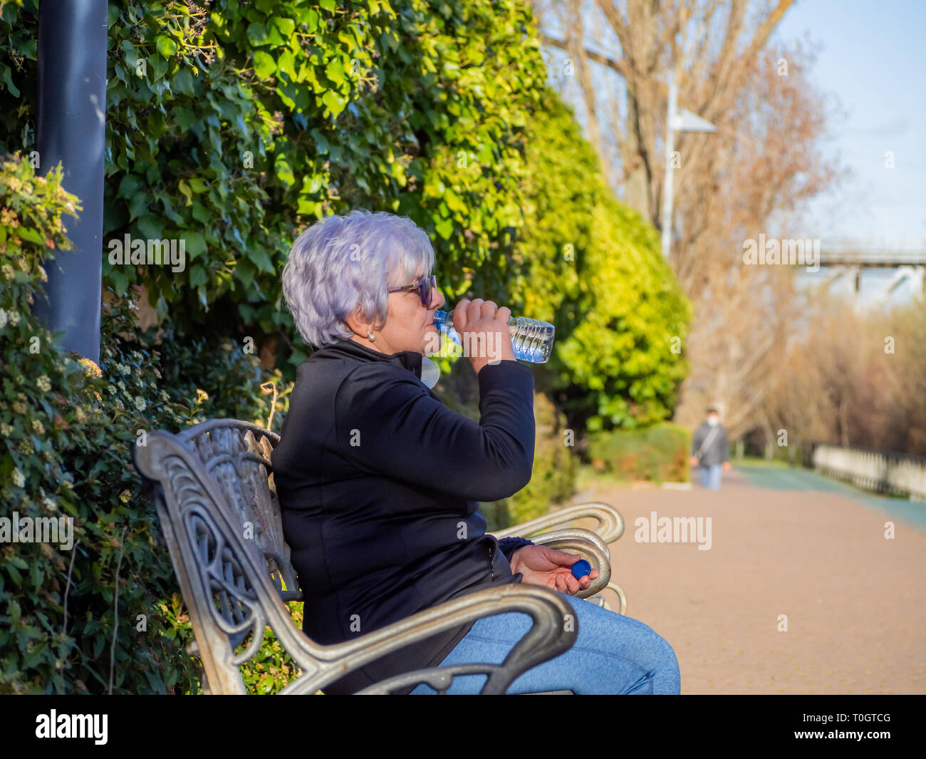 A senior woman with white hair sitting on a park bench drinking water from a plastic bottle Stock Photo