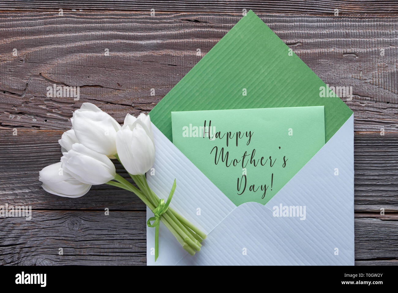 Springtime flat lay, white tulips and green card 'Happy Mother's day' in paper envelope on rustic wooden background Stock Photo