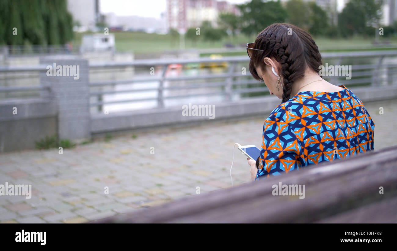 young woman sitting on a bench in the park and listening to music Stock Photo