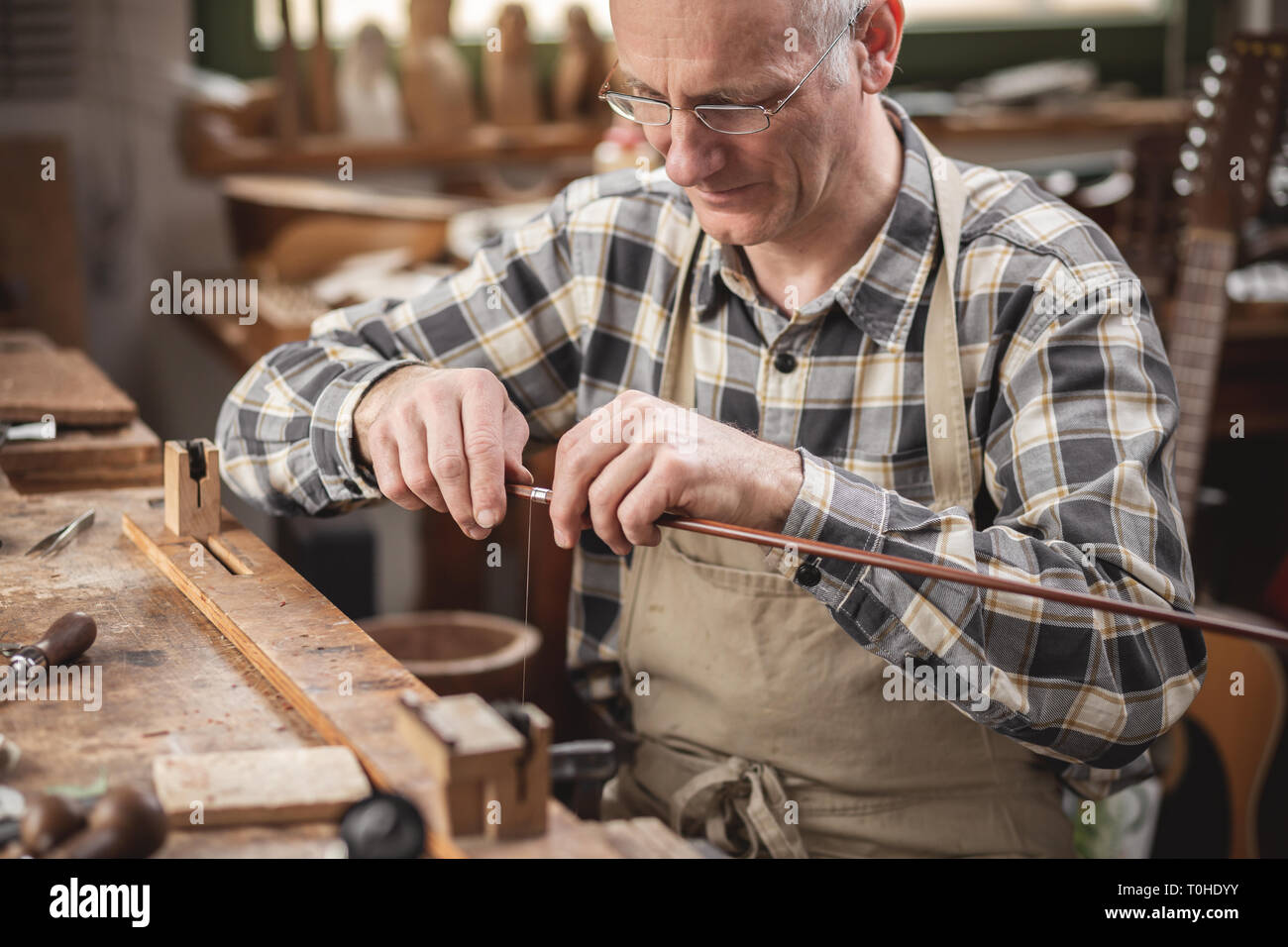 Mature instrument maker inside a rustic workshop is winding a wire around a violin bow Stock Photo