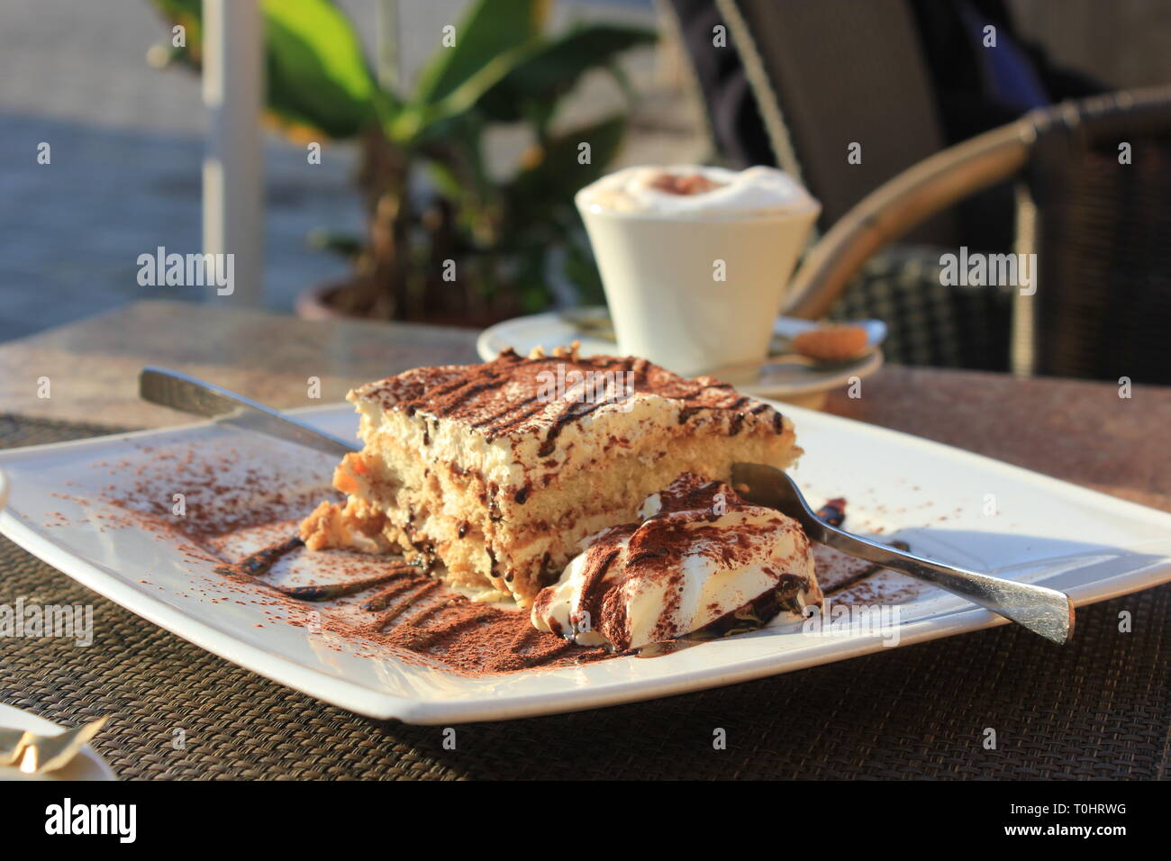 Side view on freshly made Tiramisu with cocoa powder and choco dressing, a cappuchino cup in background. One for two, cake with two forks. Stock Photo