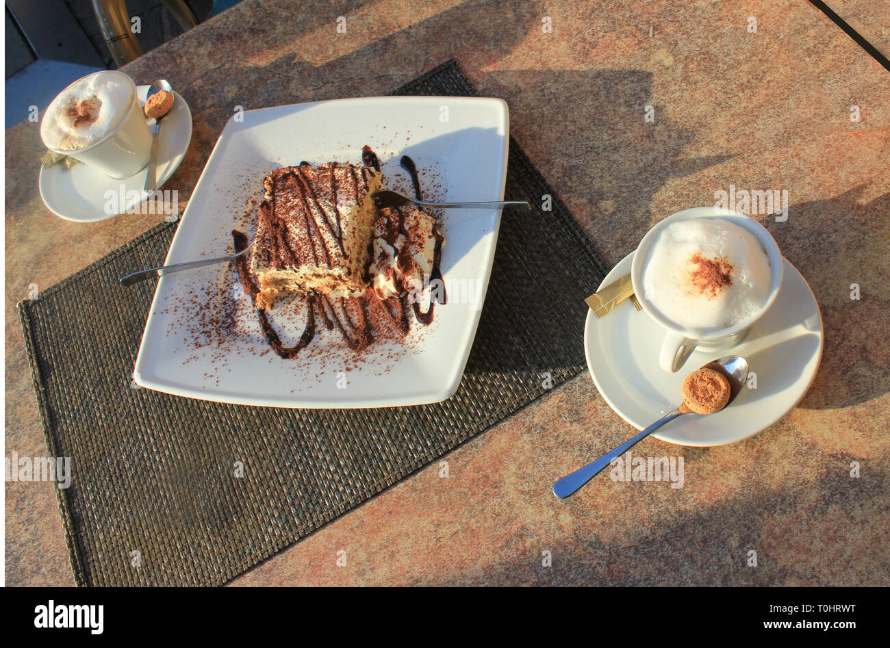 Freshly made Tiramisu with cocoa powder and choco dressing on a plate. Cake for two, with two forks and two fresh cappuchino cups.. Stock Photo