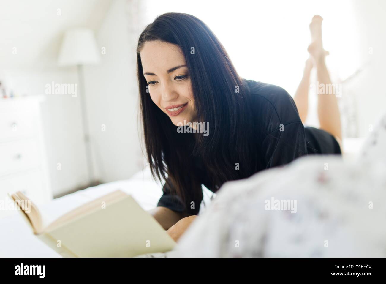 Attractive woman with dark hairs laying on bed in the morning and reading book - legs in background. Stock Photo