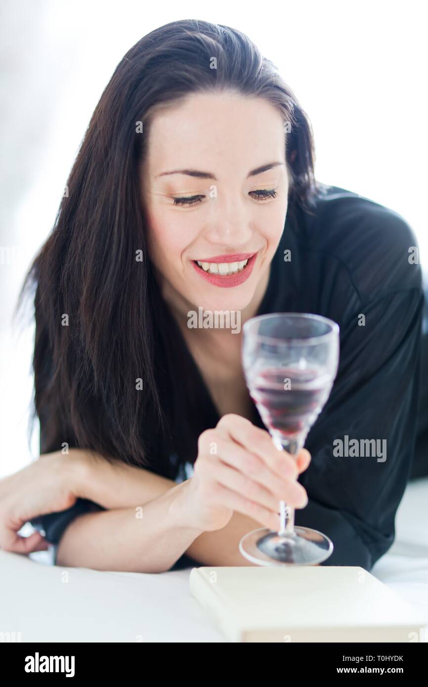 Pretty woman in black laying on bed and looking into glass of wine. Stock Photo