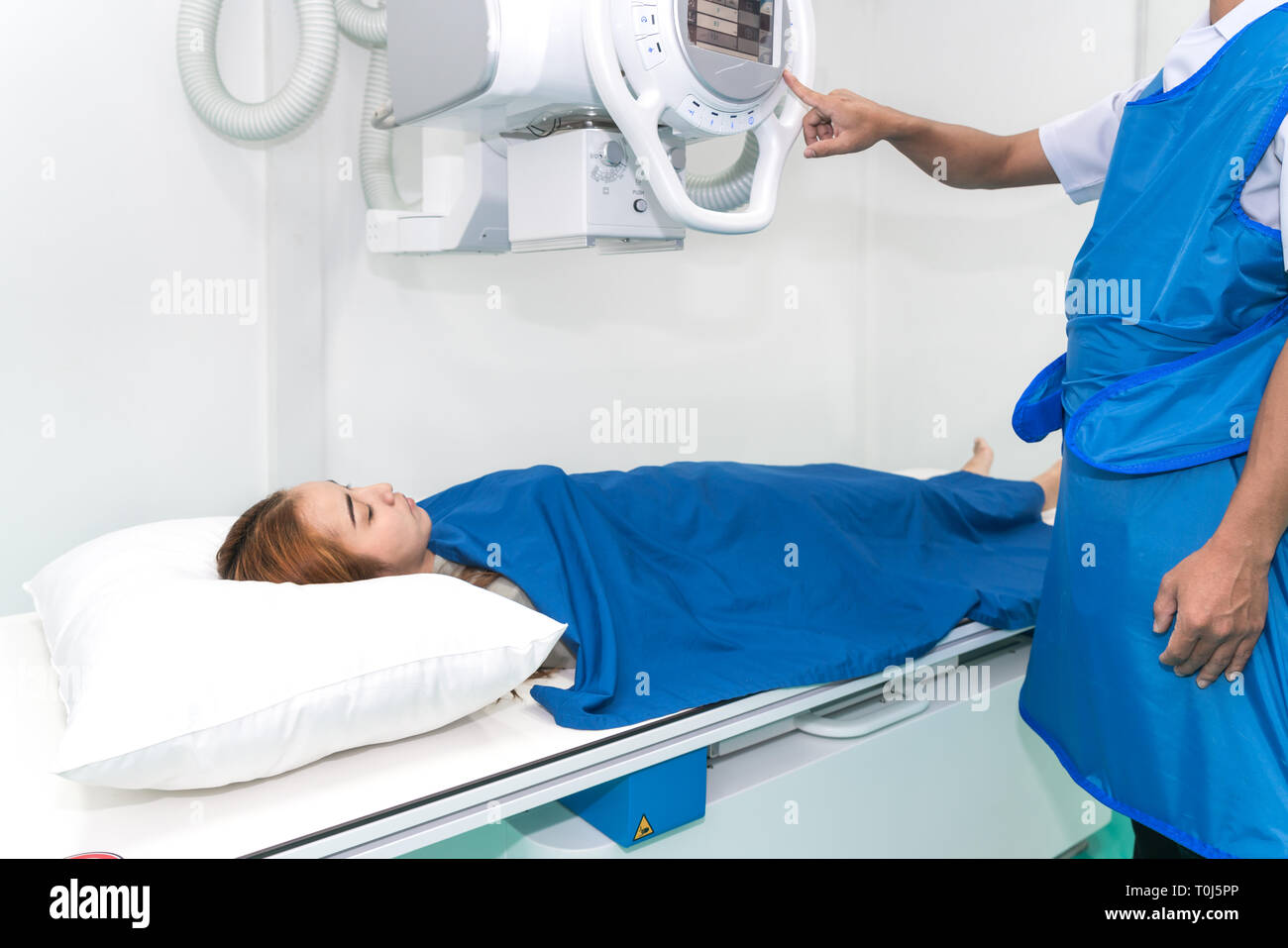 Asian young woman patient lying under X-ray device with radiologist preparing machine in X-ray room at hospital. Stock Photo