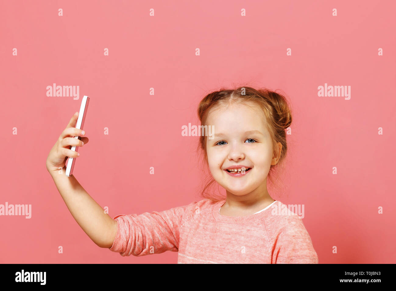 Closeup portrait of a cute little girl with buns of hair on a pink background. The child holds the phone and takes a selfie Stock Photo