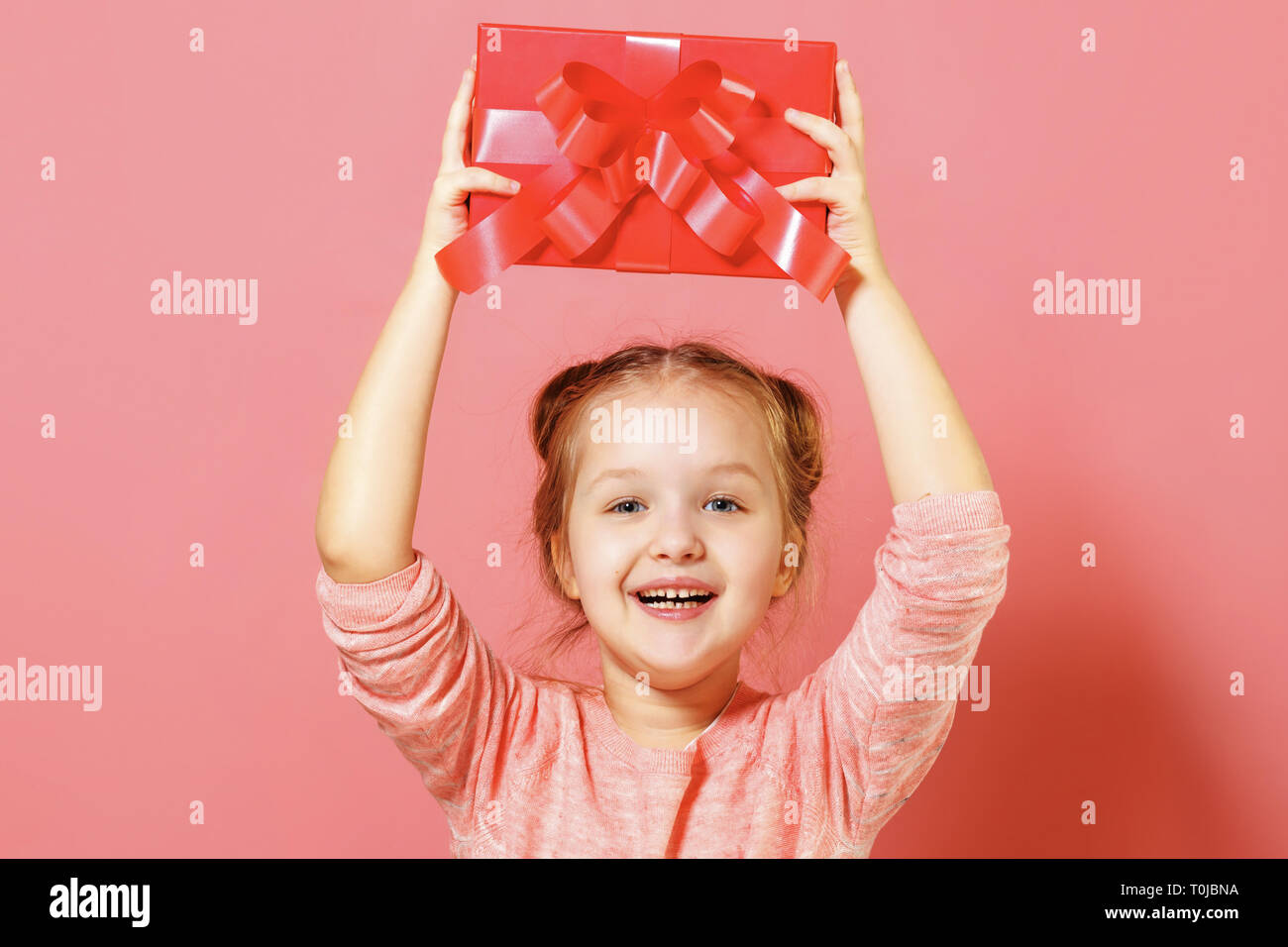 Closeup portrait of a cute little girl with hair buns on pink background. A child above his head holds a box with a gift. Stock Photo
