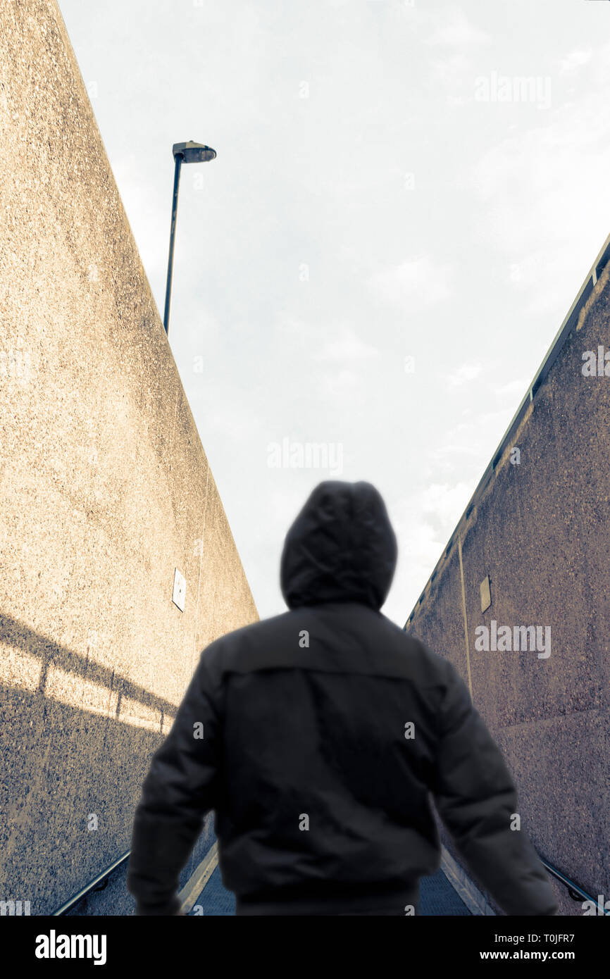 Rear view of a silhouetted figure in a hoodie, back to camera, emerging from an underpass and walking away on an upward slope Stock Photo