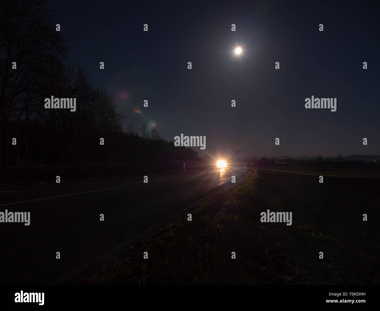 Winding Country Road Illuminated by the Headlights of an Approaching Car with Full Moon and Mysterious Landscape Stock Photo