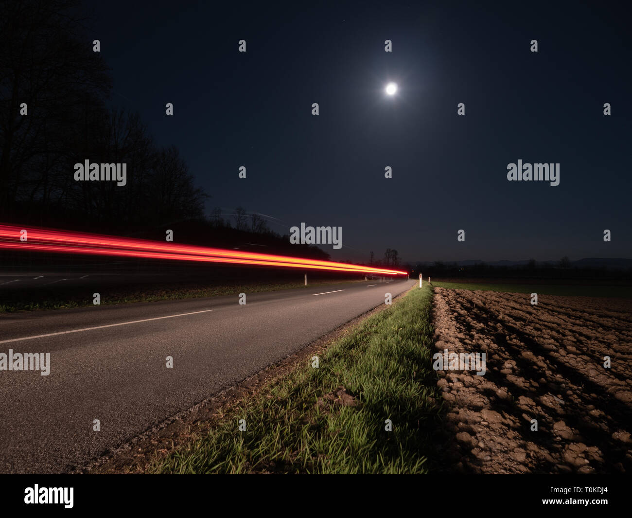 Country Road at Night Illuminated by a Passing Car  with Full Moon and Red Light Trails - Long Exposure Stock Photo