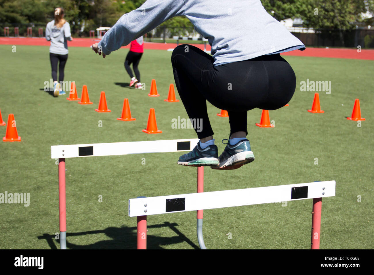 High school girls working out on a turf field by jumping over hurdles and doing running drills over orange cones. Stock Photo