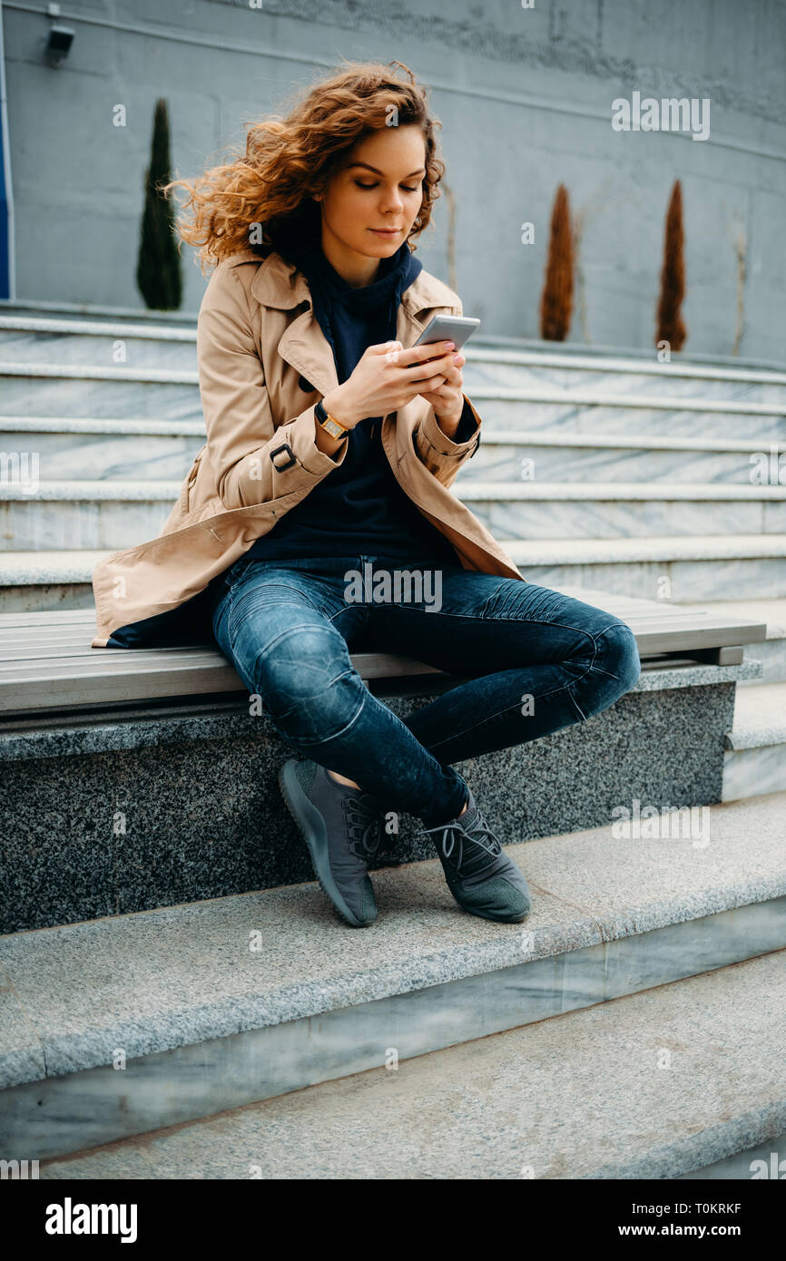 Portrait young woman wearing beige trench coat and blue jeans sitting on bench crossing legs using smart phone. Female holding mobile device and typin Stock Photo