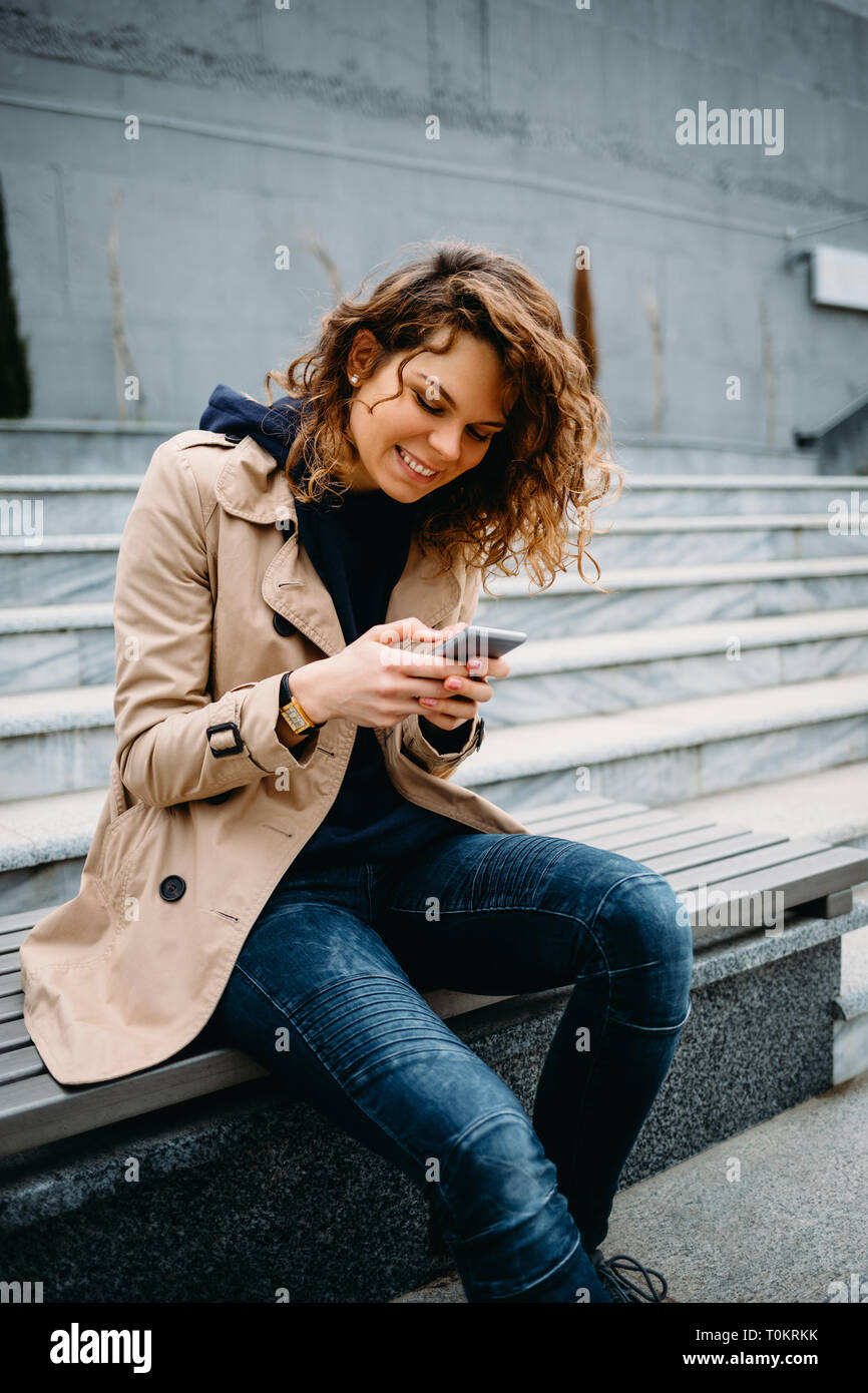 Portrait smiling young woman wearing beige trench coat and blue jeans sitting on bench using smart phone. Happy female holding mobile device and typin Stock Photo