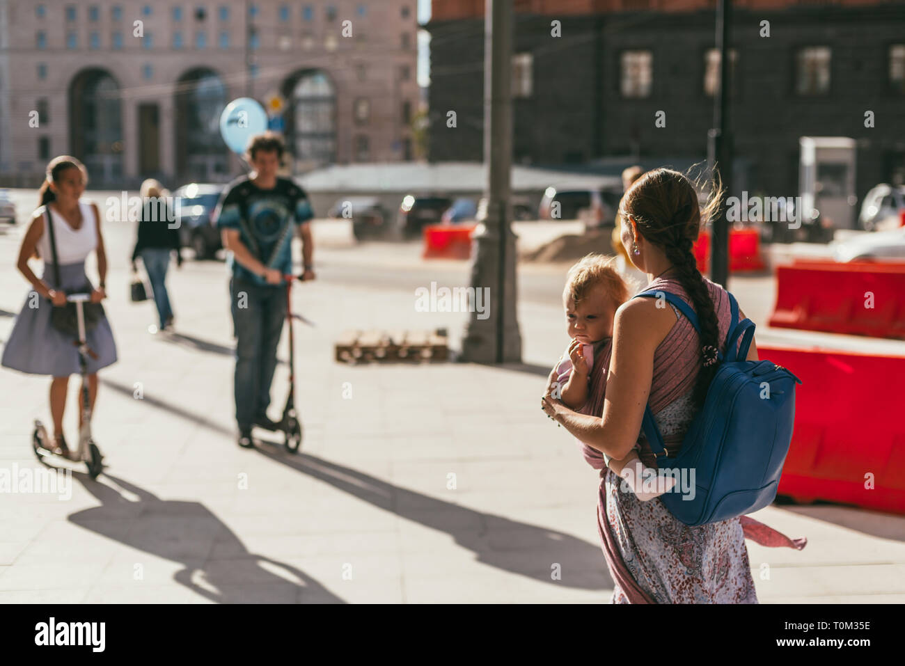 MOSCOW, RUSSIA - AUGUST 14, 2015. Young mother with a baby in a sling in the front and a backpack behind her back walking in the city. A young boy and Stock Photo